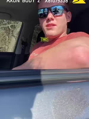 A man wearing sunglasses is inside of a car with the windows down. The image is dominated by his upper body and face, with the car interior visible in the background.
