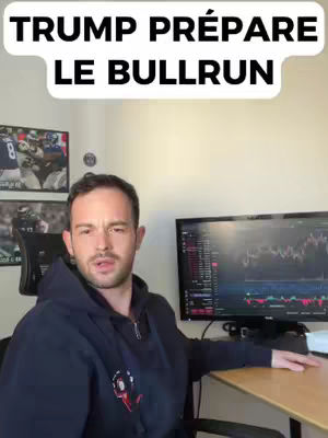 A man in a dark blue hoodie sits at a desk, looking towards the camera. Above him on the screen is the text 'Trump Prépare le Bullrun' in large white font within a white rounded rectangle overlay. Behind him are framed images and a computer monitor displaying a line graph.