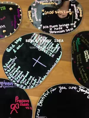 The image shows multiple round black surfaces with white text and drawings on them, arranged on a light-colored wooden surface. In the middle of the image, there's a person visible, reflected in one of the black circles. The text includes phrases such as 'God says I am...' and other religious sentiments. The style is simple, handwritten, and appears to be used for a bible study idea.