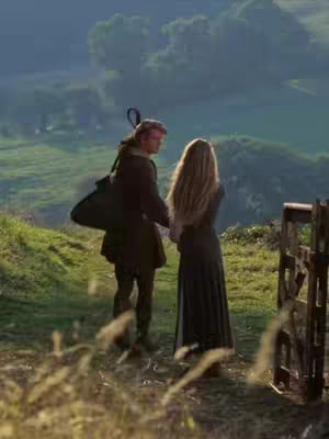 A couple stands facing away from the camera on a grassy hilltop, holding hands. The man, on the left, wears a dark coat with a bag slung over his shoulder. The woman, on the right, has long flowing hair and is wearing a long dark dress. They are looking out over a green landscape with a few clouds in the sky. There is a rustic wooden fence to the right. The overall aesthetic is reminiscent of a medieval or fantasy setting.