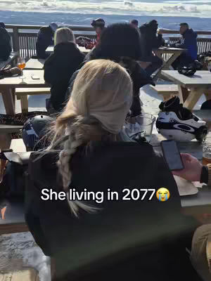 A blonde woman with a braid is seen from the back, seated at a table outside with other people visible in the background. The scene is outdoors with a snowy area and a view of the sky. Text overlaid on the image reads, "She living in 2077". The overall composition suggests a candid or observational style.