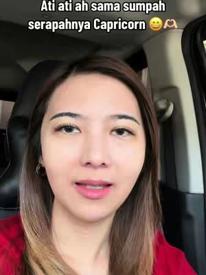 A woman with light skin and long, ombre-brown hair is centered in a selfie-style video. She is wearing a red shirt. The background is blurred, showing a car interior. Above her head, text in Indonesian reads, 'Be careful with the oath of how Capricorn is', followed by smiley face emojis and hands making the 'ok' sign.