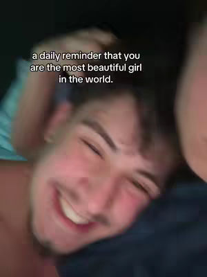 A close-up shot of a man's face, partially obscured by another person's arm and shoulder. The man is smiling with his eyes closed, indicating happiness or contentment. Text overlayed on the image reads, 'a daily reminder that you are the most beautiful girl in the world'. The background is dark, suggesting a dimly lit environment.