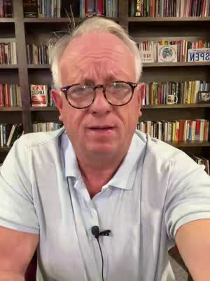 A man with glasses stands in front of a bookshelf, looking at the camera. He is wearing a light blue polo shirt. The background consists of bookshelves filled with books and some decor.