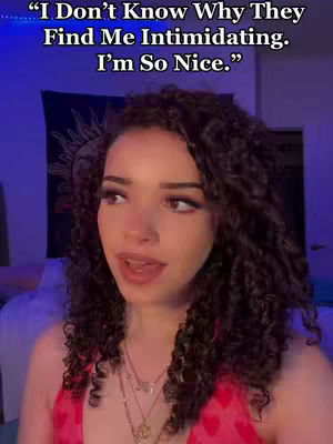 A young woman with curly brown hair and a pink top is the central figure. She has makeup on and is looking slightly off-camera. Text is overlaid at the top of the frame, and the blurred background suggests an indoor setting, possibly a bedroom with a tapestry visible.