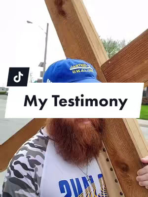 Image features a man carrying a wooden cross on his shoulders, with his head and upper body visible. He wears a baseball cap, a t-shirt, and has a beard. Text overlay reads 'My Testimony'. The background is blurred but appears to be a street or outdoor setting.