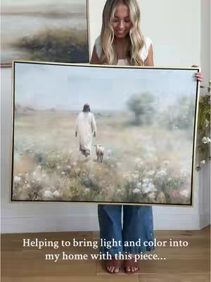 A woman holds a framed art print of Jesus walking with a lamb in a field. She is smiling at the camera.