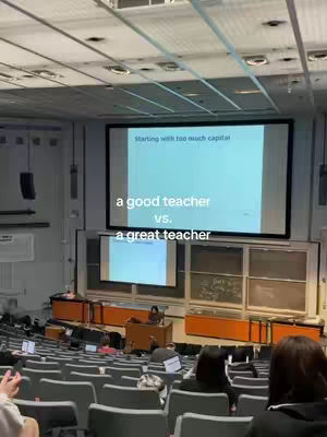 The image shows a large lecture hall with rows of seats facing a stage. On the stage, there's a presenter in front of a screen displaying text. The text overlay reads, 'a good teacher vs. a great teacher'.