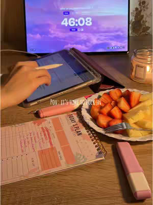 A wooden table is the setting for a study session. On the left, an open notebook with pink and peach color-coded sections. The right side holds a plate of cut strawberries and what appears to be pineapple. A pink highlighter and a pen are also present. In the background sits a tablet with a stylus, and a computer monitor displays a timer. The color palette is soft, with natural wood tones and a focus on light pink and peach hues.