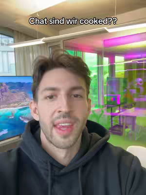 A man with short brown hair, wearing a black hoodie, is positioned in front of a blurred office environment, with a large television screen displaying a landscape behind him. The background features various colored glass or panels, and the lighting is a mix of indoor fluorescent and natural light. Above the man's head is overlay text.