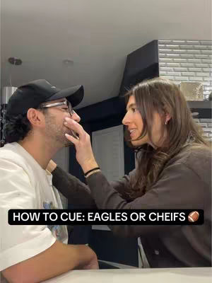 A woman holding her hands on a man's face. The man is wearing a hat and glasses, and the woman is smiling.