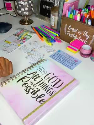 A neatly organized desk with a variety of items related to journaling and prayer. The centerpiece is a spiral-bound journal with the words "WITH GOD ALL THINGS ARE POSSIBLE" in large, elegant script. Around the journal, there are colorful pens, sticky notes, stickers, and decorative tape. In the background, there's a lit candle, a sign that says "Grateful spirit", and other office supplies.