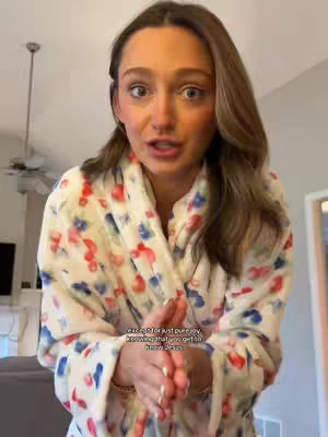A woman with light skin and long brown hair is centered in the frame, wearing a white robe with a playful floral pattern of red and blue. She is speaking, likely directly to the camera, her facial expressions suggesting earnest communication or sharing of personal feelings. She is standing in what appears to be a residential living space, with a light fixture and ceiling in the background adding context to the setting.