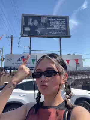 A woman with black and silver hair is posing in front of a billboard. She is wearing sunglasses and has her hands up with a pointing gesture.