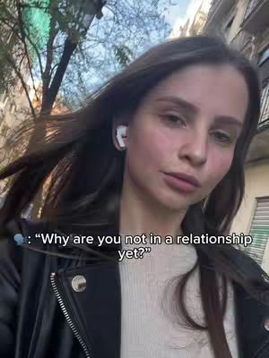 A woman with long brown hair stands outside with buildings in the background. She is wearing a black leather jacket and a white top. Text overlayed on the video reads, "Why are you not in a relationship yet?"