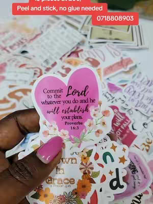 The image shows stickers of various shapes and sizes, with a hand holding a heart-shaped sticker with a bible verse. The stickers are in various colors and designs. The background is cluttered with other stickers.