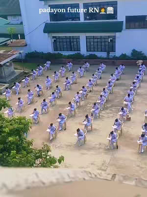 An aerial shot of a courtyard filled with rows of students seated at individual desks. They are all facing the same direction and appear to be taking an exam. The background features a large building with several windows and a green awning. The sky is overcast and the overall composition is simple and organized.