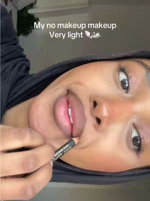 A close-up shot of a person applying lip liner. The person is facing slightly to the left, with their mouth open and a pencil in hand. The background is blurred, suggesting a focus on the subject.