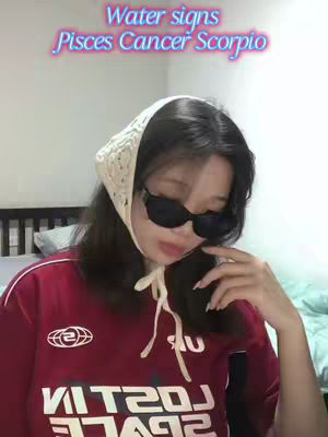 A young woman with long dark hair is wearing a cream-colored headscarf and dark sunglasses. She wears a red shirt with a white design across the chest. The text over the image reads 'Water signs Pisces Cancer Scorpio' in a playful font with a neon effect.