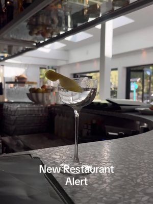 The image shows a cocktail glass with a clear liquid and a lemon twist, placed on a bar counter. The background is a restaurant setting, with open, bright spaces and what appears to be a window or skylight.