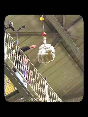 A photograph featuring a person standing on a metal staircase, with a contraption hanging. The metal staircase has white railings with a structure above. A person's arm is extended, possibly manipulating the contraption. The composition is set against the off-white background, creating depth.