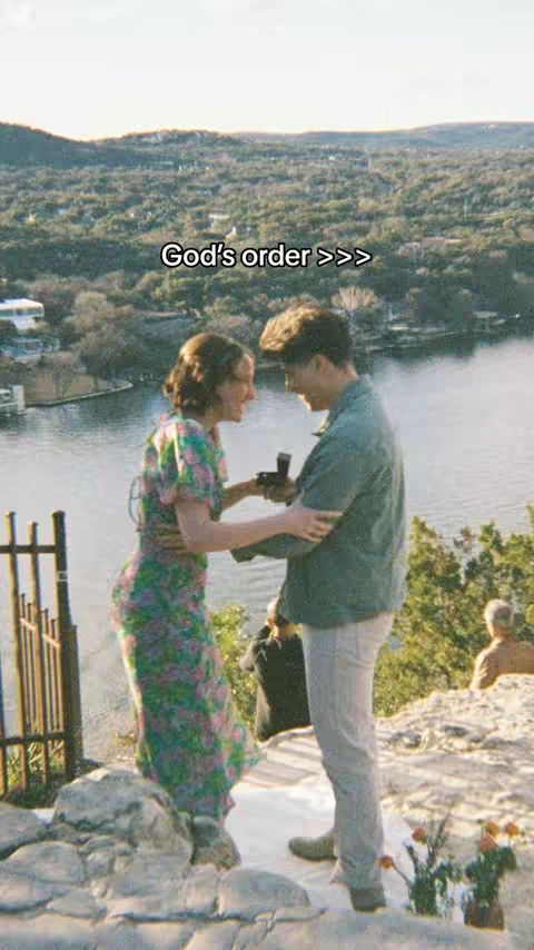 The image portrays a proposal scene on a scenic cliffside overlooking a vast lake and green landscape. A woman in a floral midi dress stands facing a man, mid-embrace, during the proposal. The man in a button down shows her what appears to be a ring box as she excitedly engages with the moment, the background suggesting an outdoor celebration of their engagement.