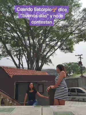 Two women are standing outdoors in front of a building and a large tree with an overlay of text. One woman is wearing a black top and jeans. The other woman is in a black and white striped dress. The sky is overcast.