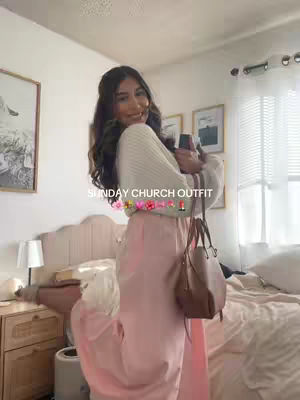 A young woman stands in a bedroom, dressed in a modest, light-colored outfit. She smiles and holds a phone, suggesting a photo or video shoot. The room has a bed with a white headboard and curtains. The video overlayed in the background says 'SUNDAY CHURCH OUTFIT'. Overall, the image presents a fashion-focused, lifestyle theme.