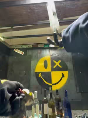 The image shows a person with black gloves trying to juggle glass bottles. The bottles, some empty, are lined up on a surface. One bottle appears to be in mid-air as if being juggled. A large smiley face is painted on the wall behind the person. The color palette is muted with some bright yellow from the smiley face.