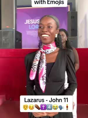 A woman with dark hair, wearing a black top and a scarf, stands in front of a purple backdrop. The woman is smiling, and a text overlay at the bottom reads, 'Lazarus - John 11' followed by various emojis. Another woman is partially visible in the background.