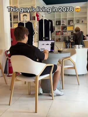 A person is seated at a table in a cafe, facing away from the camera and working on a laptop. The background features shelves with various items, and there are other people present. The color palette is neutral with some pops of color.