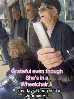 The image shows a scene of a group of people outdoors. In the foreground, a woman in a wheelchair covers her face with both hands, appearing emotional. A man stands next to her, holding a phone aloft and looking upwards, seemingly mid-speech or explanation. Behind them are two more men. The setting looks urban, with a building or shopfront behind them. The image has text overlays in a pink hue. The overall composition suggests a moment of shared experience or possible explanation in a social context outside.