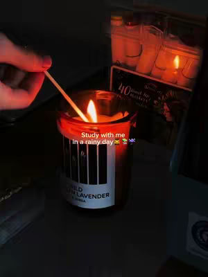 The cover image shows a candle lit in a glass holder, the flame is bright. A hand, with painted fingernails, holds a match near the wick. Overlayed text reads 'Study with me in a rainy day'. The overall aesthetic is warm and cozy.