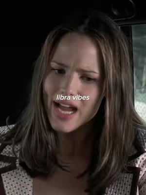 A medium shot of a woman with long brown hair. She is looking to the right and speaking with an open mouth, a text overlay "libra vibes" in the center. The background appears to be a neutral, possibly indoor setting.