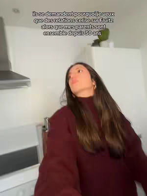 A young woman in a maroon sweater is positioned slightly off-center in a kitchen setting. She's looking upwards and slightly to her left, with a partially visible stainless steel range hood behind her. The background includes a white wall and some green plants, suggesting a home environment. Text overlays at the top of the image in French.