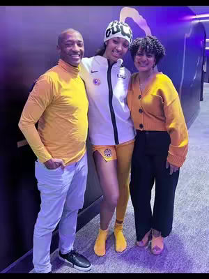 A group of three people posing for a photo, likely at a basketball game. The person in the middle is wearing a sports uniform, flanked by two people dressed casually. The background has a dark, blurred effect, and the overall composition is a mix of portraiture and candid moments.