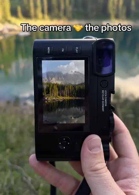 A hand holding a black camera, with the screen displaying a scenic landscape of a lake and mountains, with the hook on top.