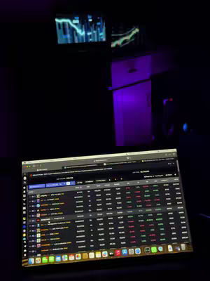 A dark room with a laptop displaying a trading interface, two monitors showing graphs, and a purple-lit wall. The composition emphasizes the technology-focused content.