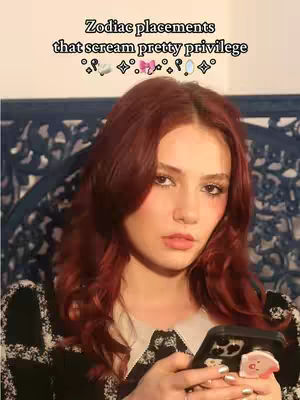 A young woman with red hair is centered in the frame, holding a phone with a pink accessory attached. The background is a patterned blue and white design. Text overlayed at the top says, "Zodiac placements that scream pretty privilege."