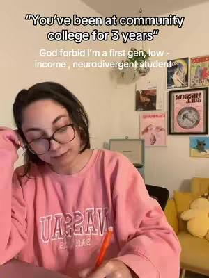 A young woman with dark hair and glasses is sitting at a desk, looking down while writing with an orange pen. She's wearing a pink sweatshirt that says "UARRAV" across the front. Behind her is a wall decorated with framed art and posters. A yellow pillow and stuffed animal sit on a yellow chair in the background.