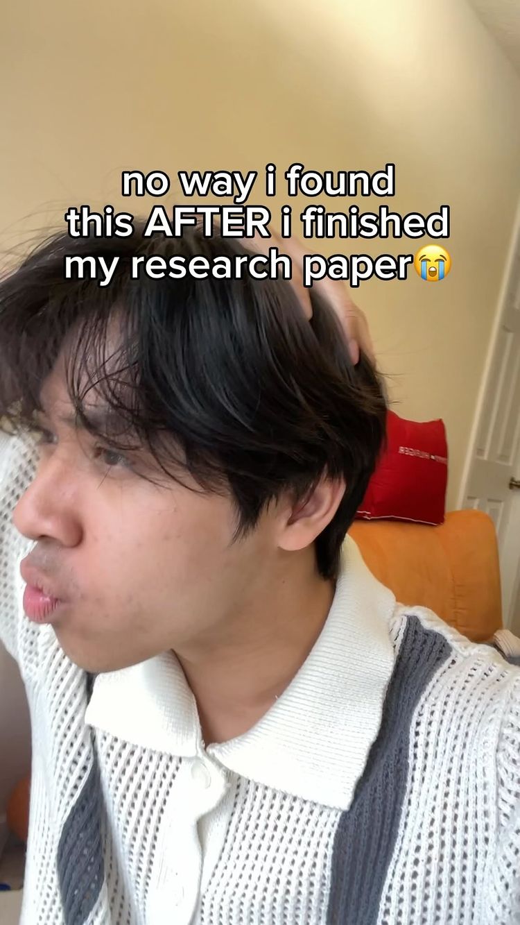 A young man with dark hair is looking at the camera with a slightly pursed expression on his lips, as if he is about to kiss. He is wearing a white sweater and has his hand in his hair. Above his head is the text overlay that reads: "no way i found this AFTER i finished my research paper". Behind him are items such as a red pillow on an orange couch. The lighting appears soft and natural.