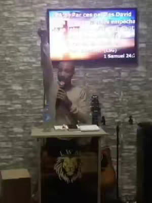 A man is standing at a podium, raising one arm in the air, with a microphone in his other hand. Behind him is a large screen displaying text and imagery related to a sermon. The background consists of a textured brick wall.