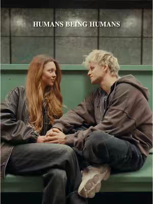 A cover image shows a couple sitting close together on a green bench, with the man looking at the woman. The woman has long reddish hair and is also looking at him. They are holding hands, suggesting an intimate moment. The background is a wall with a grid pattern and a neon sign. The overall composition emphasizes the interaction between the two figures. The image is well-lit with balanced colors.