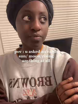 A person with dark skin wears a black head covering and a light-colored shirt with visible text. They are looking off to the side with a contemplative expression. The background includes blinds and a desk.