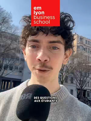 A young man with dark, curly hair and a thin mustache is holding a black microphone in front of a background of buildings. Above his head is a red square with the words 'em lyon business school' in white.