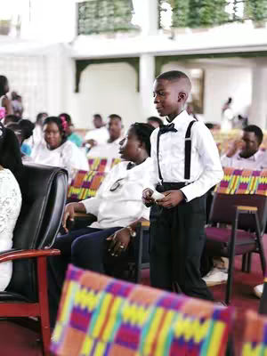 A young boy in a white shirt, black pants, suspenders, and a bow tie stands in what appears to be a church. Seated members of the congregation are softly blurred in the background.
