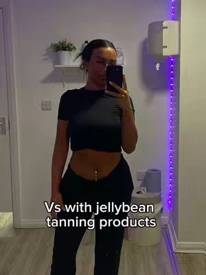 A woman in a black crop top and black pants is taking a mirror selfie in what appears to be a bathroom. The space has a white wall, a plant shelf, and purple LED lights running along the right side.