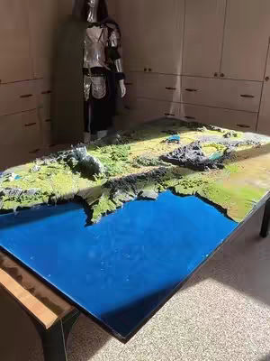 A diorama of a detailed landscape, possibly Middle-earth from 'Lord of the Rings,' is displayed on a large, rectangular table with a glossy blue surface representing water. The terrain is textured with green and brown elements, and the table is situated in a well-lit room with light-colored cabinets visible in the background. A mannequin wearing armor is in the background.