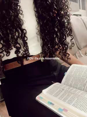 The image shows a person with long, dark curly hair. The person is wearing a white top and black bottoms, and appears to be seated with a book open. The text overlay in Portuguese says "POV: Ela é da Assembleia".