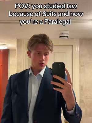 A man in a dark suit is taking a selfie in a bathroom. The background is a lit-up mirror and what appears to be a cream-colored wall. The text overlay reads 'POV: you studied law because of Suits and now you're a Paralegal'. The individual holds a black phone in his hand.